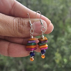 May include: A pair of dangle earrings featuring stacked, flat, rectangular beads in shades of orange, purple, green, and magenta. Each earring is finished with a dark blue faceted bead and a small, clear orange crystal at the bottom, suspended from silver-toned metal hooks.