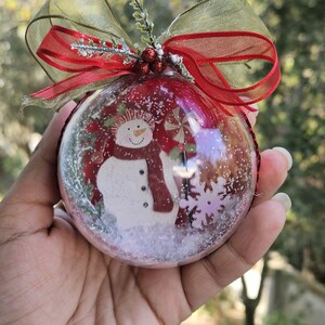 May include: A red glass Christmas ornament with a snowman inside. The snowman is wearing a red scarf and hat. The ornament is decorated with glitter and snowflakes.
