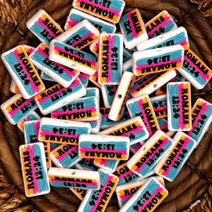 May include: A basket filled with colorful rectangular plastic charms with the text "Romans 12:24" printed on them. The charms have a blue, orange, and pink color scheme.