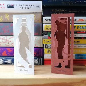 Pode incluir: Dois marcadores de página de papel com o tema Bons Preságios. O marcador da esquerda é branco com uma silhueta de um homem de terno. O marcador da direita é marrom com uma silhueta de uma cobra e um homem.