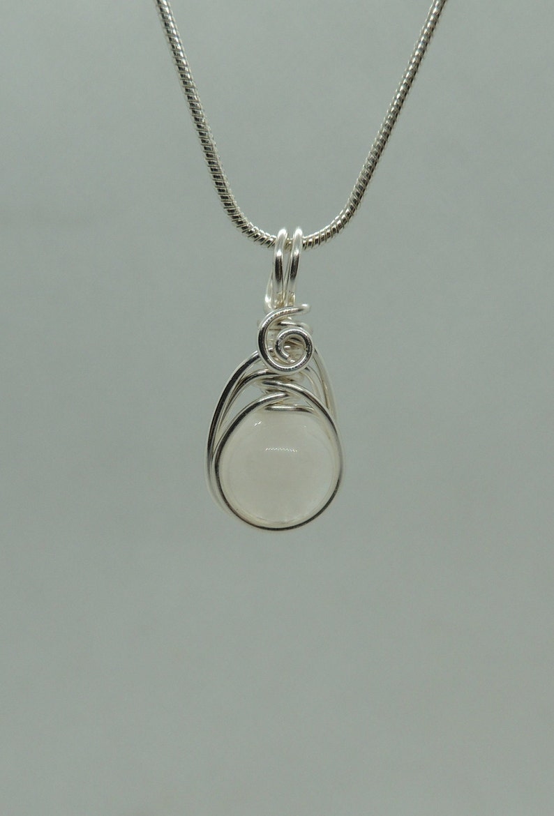 Puede incluir: Un colgante de alambre plateado que envuelve una piedra blanca opaca, lisa y redonda. El alambre de metal forma una espiral decorativa en la parte superior y asegura la piedra dentro de una estructura tipo jaula. El colgante cuelga de una cadena.