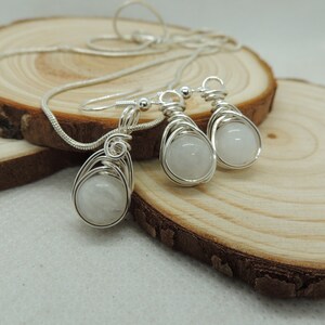 Puede incluir: Un conjunto de joyer&iacute;a compuesto por un collar y dos pendientes a juego de color plateado. Cada pieza presenta una piedra preciosa redonda de color blanco lechoso envuelta en un dise&ntilde;o de alambre decorativo de color plateado.