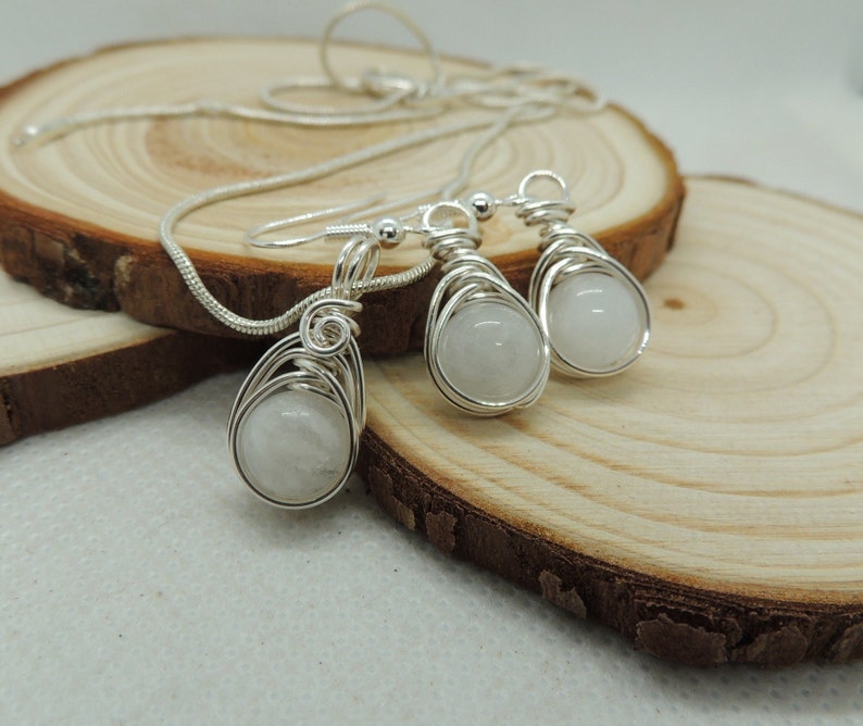 Puede incluir: Un conjunto de joyer&iacute;a compuesto por un collar y dos pendientes a juego de color plateado. Cada pieza presenta una piedra preciosa redonda de color blanco lechoso envuelta en un dise&ntilde;o de alambre decorativo de color plateado.