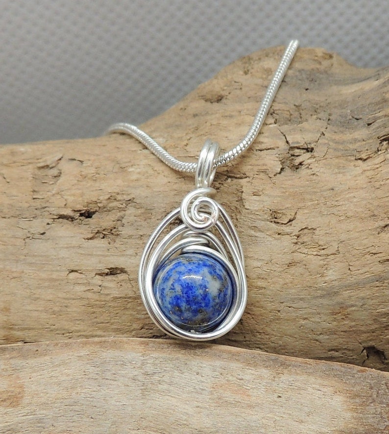 Puede incluir: Un colgante redondo de lapisl&aacute;zuli azul profundo con motas doradas, encerrado en un dise&ntilde;o de alambre plateado. El colgante cuelga de una cadena de serpiente plateada a juego, que presenta un detalle de alambre en espiral en la parte superior.