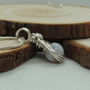 May include: A silver-toned pendant featuring a single round, pale lavender-grey bead encased in a decorative wire-wrapped design. The pendant is attached to a thin, flexible silver-toned snake chain that loops through a circular wire bail at the top.