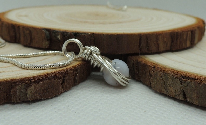 May include: A silver-toned pendant featuring a single round, pale lavender-grey bead encased in a decorative wire-wrapped design. The pendant is attached to a thin, flexible silver-toned snake chain that loops through a circular wire bail at the top.
