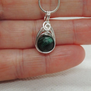 Puede incluir: Un colgante con una piedra redonda de color verde oscuro jaspeado, envuelta en alambre de color plateado. El alambre forma un marco en forma de l&aacute;grima con un peque&ntilde;o detalle en espiral en la parte superior, colgado de una cadena fina plateada.