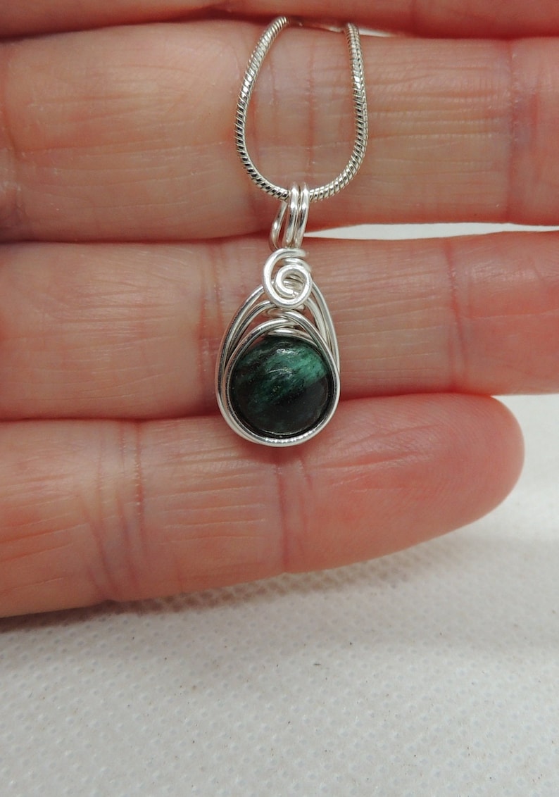 Puede incluir: Un colgante con una piedra redonda de color verde oscuro jaspeado, envuelta en alambre de color plateado. El alambre forma un marco en forma de l&aacute;grima con un peque&ntilde;o detalle en espiral en la parte superior, colgado de una cadena fina plateada.