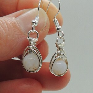 May include: A pair of silver-toned wire-wrapped drop earrings featuring round, translucent white beads with subtle brown inclusions. Each earring is suspended from a silver-toned hook and secured with a coiled wire cage design around the bead.