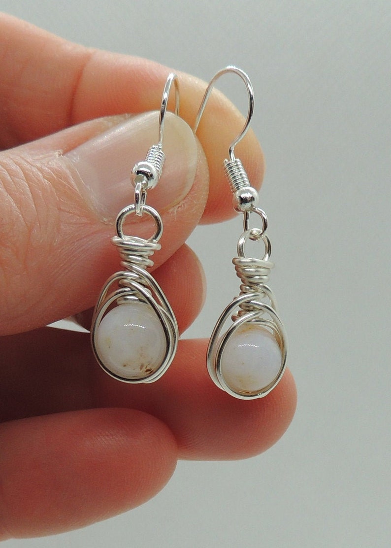 May include: A pair of silver-toned wire-wrapped drop earrings featuring round, translucent white beads with subtle brown inclusions. Each earring is suspended from a silver-toned hook and secured with a coiled wire cage design around the bead.