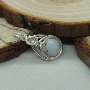 May include: A silver-toned wire-wrapped pendant featuring a smooth, round, pale blue stone with subtle grey inclusions. The pendant is attached to a matching silver-toned snake chain necklace, showcasing intricate metal coils around the stone.