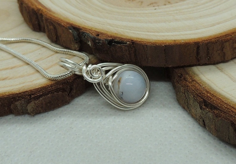 May include: A silver-toned wire-wrapped pendant featuring a smooth, round, pale blue stone with subtle grey inclusions. The pendant is attached to a matching silver-toned snake chain necklace, showcasing intricate metal coils around the stone.