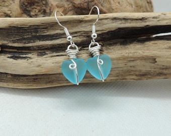 Puede incluir: Un par de pendientes colgantes con cuentas de vidrio esmerilado en forma de corazón de color azul claro. Cada cuenta está envuelta en alambre de tono plateado que forma una espiral decorativa, suspendida de ganchos de metal plateado.