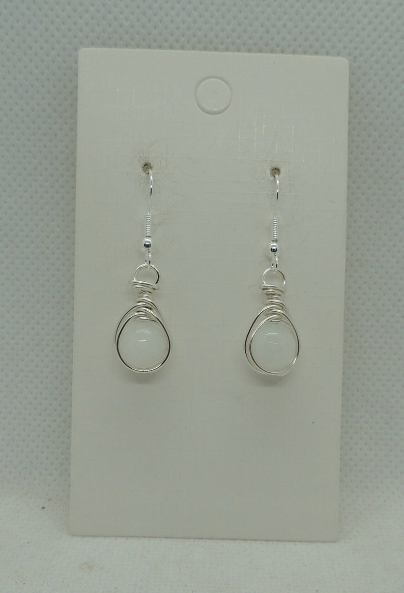 Puede incluir: Un par de pendientes colgantes de alambre plateado con cuentas redondas de color blanco opaco. Los pendientes est&aacute;n sujetos a una tarjeta de presentaci&oacute;n rectangular de color blanco con un orificio circular en la parte superior para colgar.