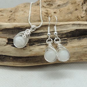 Puede incluir: Un collar con colgante de alambre de plata y un par de pendientes colgantes a juego, cada uno con una piedra preciosa blanca transl&uacute;cida, redonda y lisa, sujeta dentro de una jaula decorativa de alambre met&aacute;lico de tono plateado.