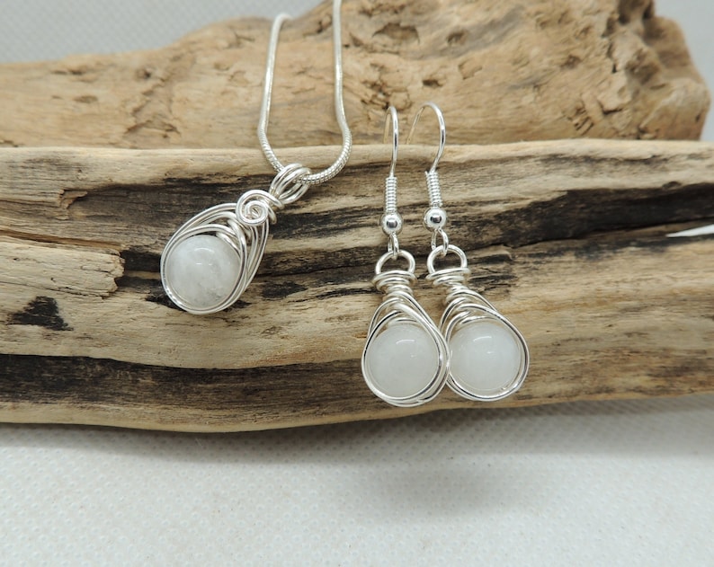 Puede incluir: Un collar con colgante de alambre de plata y un par de pendientes colgantes a juego, cada uno con una piedra preciosa blanca transl&uacute;cida, redonda y lisa, sujeta dentro de una jaula decorativa de alambre met&aacute;lico de tono plateado.