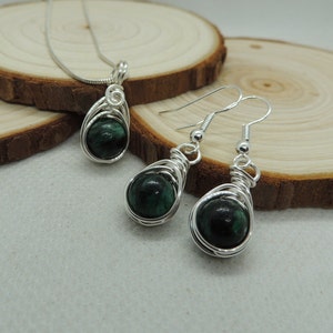 Puede incluir: Un conjunto de joyer&iacute;a que consta de un colgante en una cadena plateada y un par de pendientes a juego. Cada pieza presenta una piedra preciosa redonda, de color verde oscuro y pulida, envuelta en un dise&ntilde;o de alambre plateado con espiral.