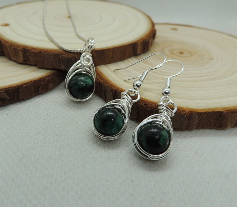 Puede incluir: Un conjunto de joyer&iacute;a que consta de un colgante en una cadena plateada y un par de pendientes a juego. Cada pieza presenta una piedra preciosa redonda, de color verde oscuro y pulida, envuelta en un dise&ntilde;o de alambre plateado con espiral.