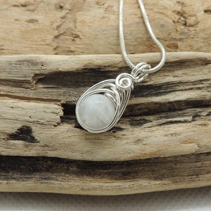 Puede incluir: Un colgante de alambre plateado con una piedra preciosa redonda y blanca transl&uacute;cida. El colgante cuelga de una cadena de serpiente plateada a juego, con el alambre formando una figura de l&aacute;grima alrededor de la piedra central.
