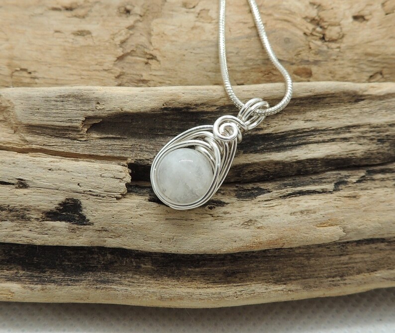 Puede incluir: Un colgante de alambre plateado con una piedra preciosa redonda y blanca transl&uacute;cida. El colgante cuelga de una cadena de serpiente plateada a juego, con el alambre formando una figura de l&aacute;grima alrededor de la piedra central.