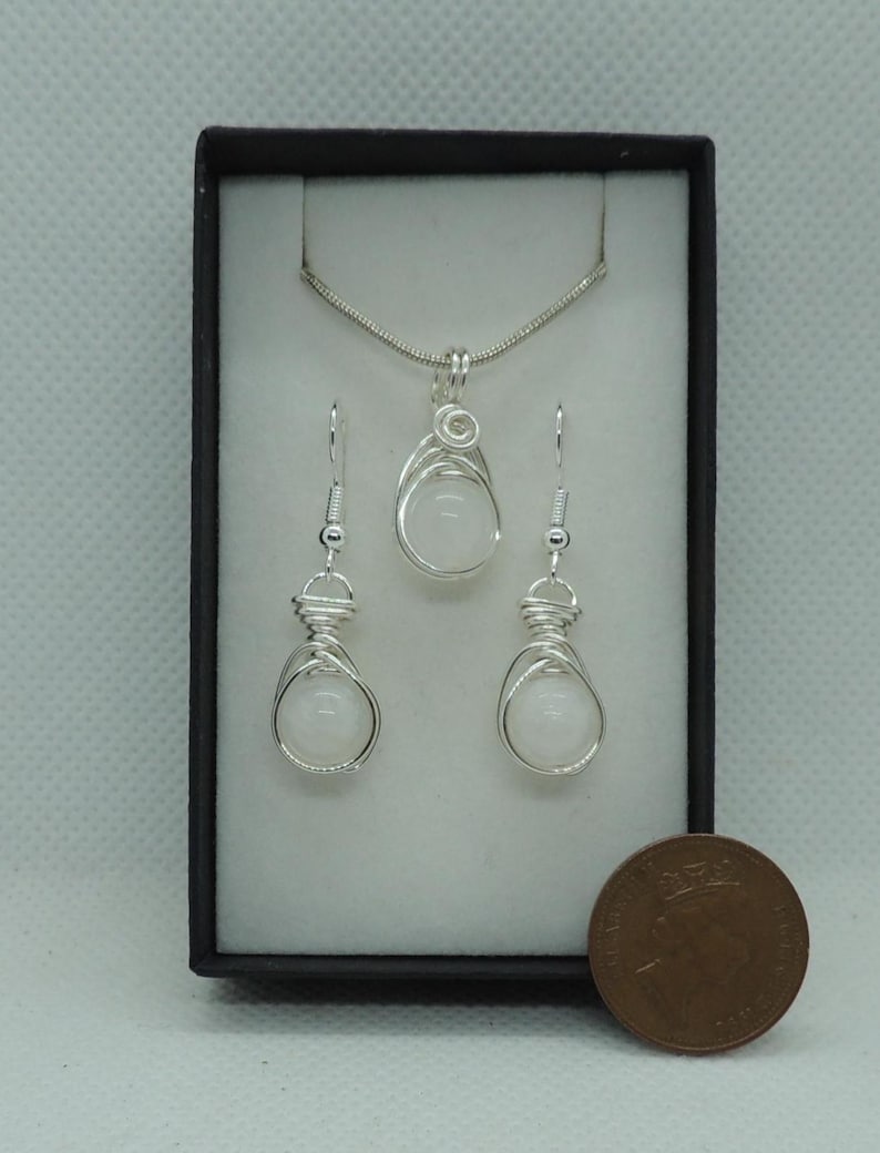 Puede incluir: Un conjunto de joyer&iacute;a que consta de un collar con colgante y dos pendientes a juego, cada uno con una piedra blanca redonda envuelta en alambre de color plateado. El conjunto se presenta en una caja negra junto a una moneda de cobre.