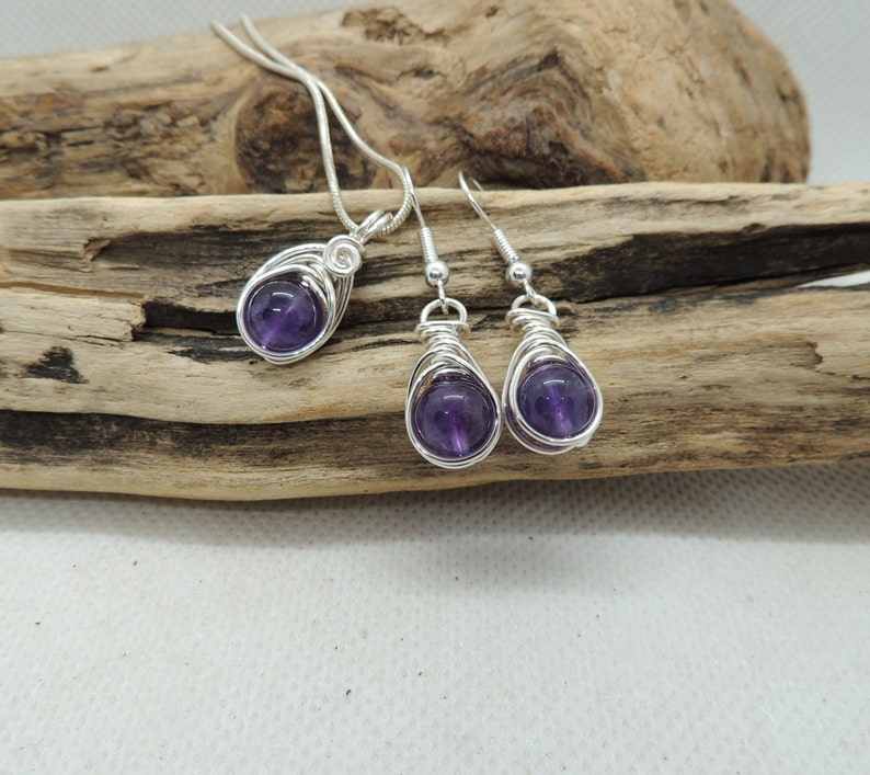 May include: A set of amethyst gemstone jewelry featuring a pendant necklace and matching earrings. The amethyst stones are wrapped in silver wire and have a deep purple hue.