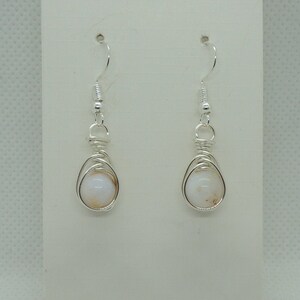 May include: A pair of silver-toned wire-wrapped earrings featuring round, opaque white beads with subtle tan inclusions. The earrings are suspended from silver-toned fishhook wires and are displayed on a rectangular white card with a circular hole.