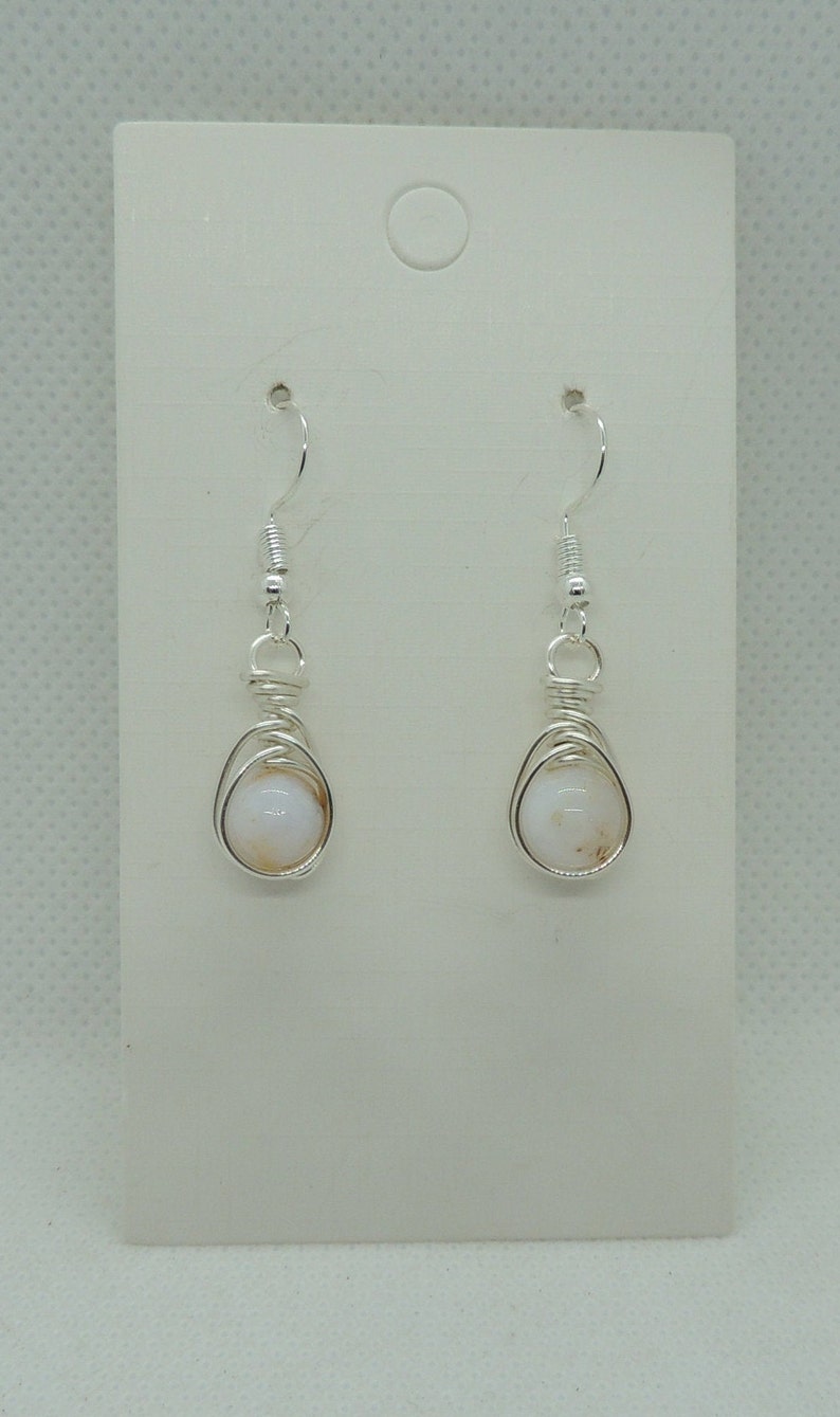 May include: A pair of silver-toned wire-wrapped earrings featuring round, opaque white beads with subtle tan inclusions. The earrings are suspended from silver-toned fishhook wires and are displayed on a rectangular white card with a circular hole.