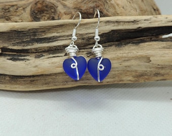 Puede incluir: Un par de pendientes con cuentas de vidrio azul intenso en forma de corazón envueltas en alambre de tono plateado. Cada pendiente cuelga de un gancho plateado simple, con el alambre formando un acento en espiral sobre el corazón azul.