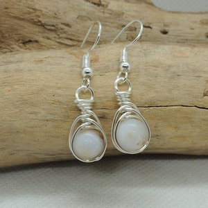 May include: A pair of silver-toned wire-wrapped earrings featuring round, opaque white beads. Each earring is suspended from a silver-toned hook and showcases a decorative coil of wire securing the bead within a cage-like structure.