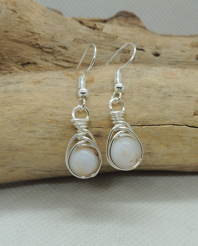 May include: A pair of silver-toned wire-wrapped earrings featuring round, opaque white beads. Each earring is suspended from a silver-toned hook and showcases a decorative coil of wire securing the bead within a cage-like structure.
