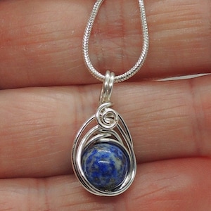 Puede incluir: Un collar con colgante que presenta una piedra preciosa de lapisl&aacute;zuli redonda de color azul intenso con motas doradas, engastada en un marco de alambre plateado con un detalle en espiral, suspendido de una cadena de serpiente plateada.