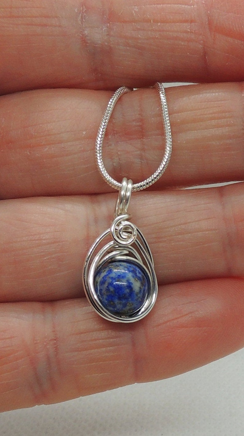 Puede incluir: Un collar con colgante que presenta una piedra preciosa de lapisl&aacute;zuli redonda de color azul intenso con motas doradas, engastada en un marco de alambre plateado con un detalle en espiral, suspendido de una cadena de serpiente plateada.