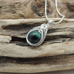 Puede incluir: Un colgante con un cabuj&oacute;n de piedra redondo de color verde oscuro y negro, engastado en una montura de alambre de color plateado. El colgante est&aacute; unido a una fina cadena de color plateado, con el alambre formando una forma de l&aacute;grima.