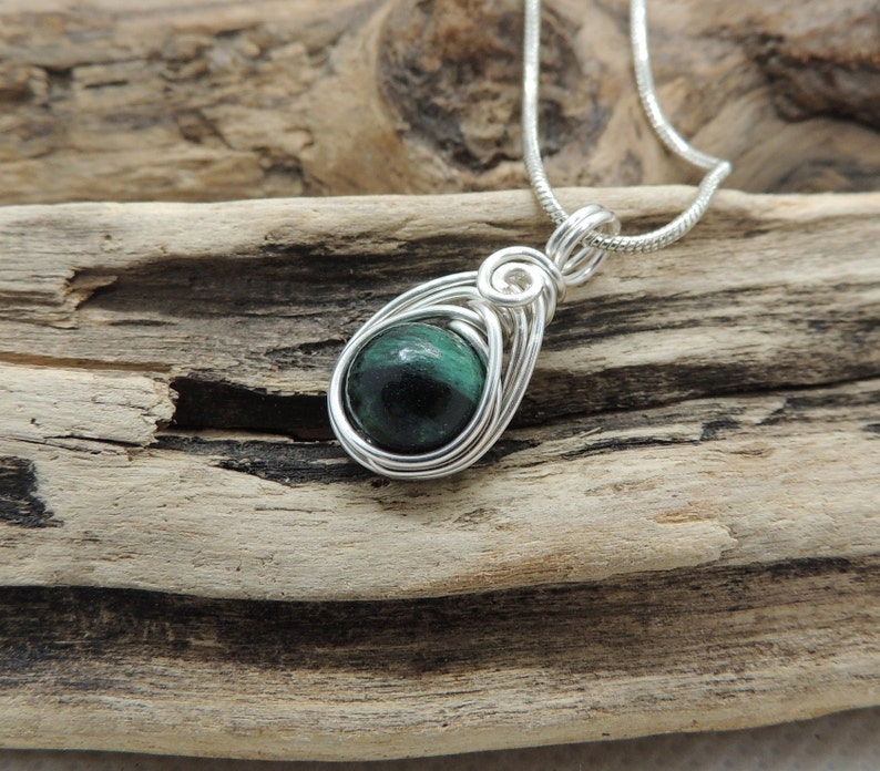 Puede incluir: Un colgante con un cabuj&oacute;n de piedra redondo de color verde oscuro y negro, engastado en una montura de alambre de color plateado. El colgante est&aacute; unido a una fina cadena de color plateado, con el alambre formando una forma de l&aacute;grima.