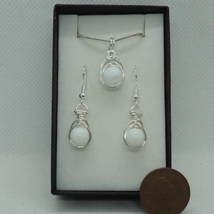 May include: A jewellery set featuring a pendant necklace and two matching drop earrings, each containing a smooth white spherical bead encased in silver-coloured wire wrapping. The set is displayed in a black box next to a copper-coloured coin.