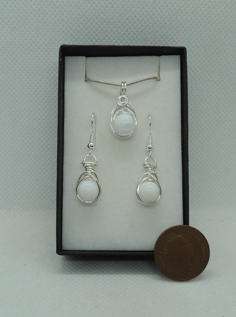 May include: A jewellery set featuring a pendant necklace and two matching drop earrings, each containing a smooth white spherical bead encased in silver-coloured wire wrapping. The set is displayed in a black box next to a copper-coloured coin.