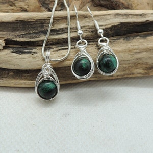 Puede incluir: Un conjunto de joyer&iacute;a que consta de un colgante en una cadena de serpiente plateada y dos pendientes a juego. Cada pieza presenta una piedra redonda veteada en verde oscuro y negro dentro de un dise&ntilde;o de alambre plateado en espiral.