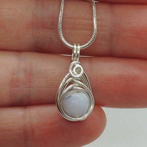 May include: A wire-wrapped pendant featuring a smooth, round, pale blue stone with subtle dark inclusions. The stone is encased in silver-toned wire that forms a teardrop-shaped frame and a small spiral detail at the top, attached to a silver snake chain.
