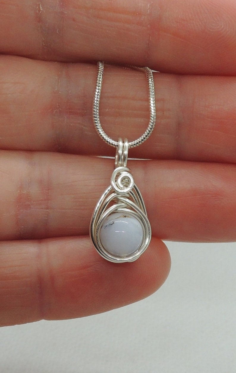 May include: A wire-wrapped pendant featuring a smooth, round, pale blue stone with subtle dark inclusions. The stone is encased in silver-toned wire that forms a teardrop-shaped frame and a small spiral detail at the top, attached to a silver snake chain.