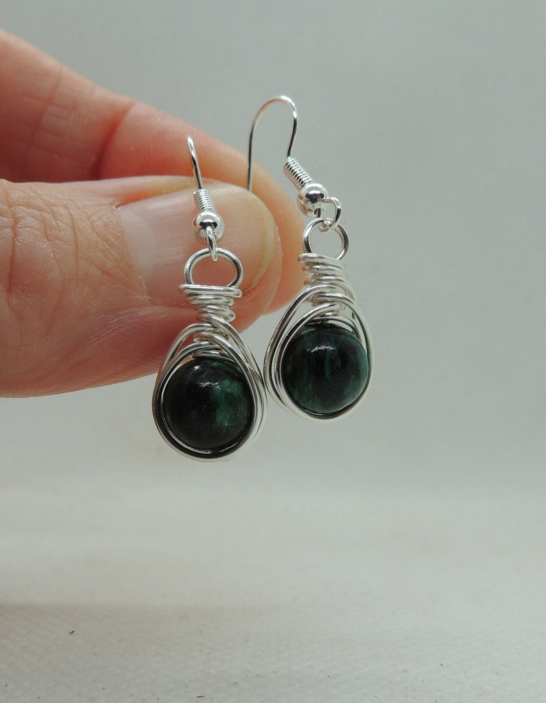 Puede incluir: Un par de pendientes colgantes con cuentas de piedra redondas, de color verde oscuro y moteadas, envueltas en alambre de tono plateado. Cada pendiente cuelga de un gancho plateado, mostrando un acabado pulido y un dise&ntilde;o de alambre enrollado alrededor de la gema.