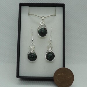 Puede incluir: Un conjunto de joyer&iacute;a que incluye un colgante y dos pendientes a juego, cada uno con una piedra esf&eacute;rica de color verde oscuro envuelta en alambre de color plateado. El conjunto se presenta en una caja negra junto a una moneda.