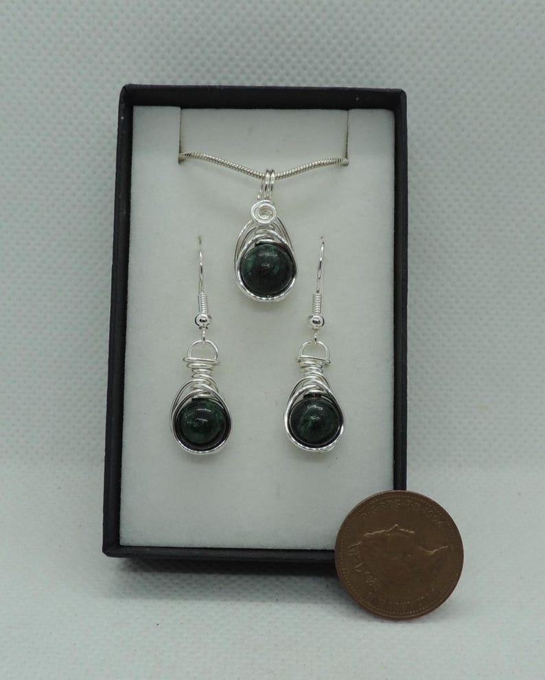 Puede incluir: Un conjunto de joyer&iacute;a que incluye un colgante y dos pendientes a juego, cada uno con una piedra esf&eacute;rica de color verde oscuro envuelta en alambre de color plateado. El conjunto se presenta en una caja negra junto a una moneda.