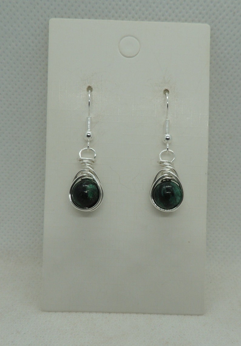 Puede incluir: Un par de pendientes colgantes con cuentas redondas de piedra verde oscuro jaspeado envueltas en alambre de tono plateado. Los pendientes est&aacute;n sujetos a ganchos de metal plateado y se presentan en una tarjeta blanca rectangular con un orificio.