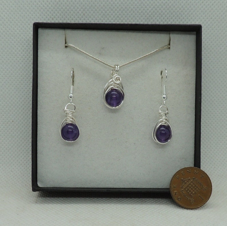 May include: A silver necklace and earrings set with amethyst stones wrapped in silver wire. The set is displayed in a black box with a British penny for scale.