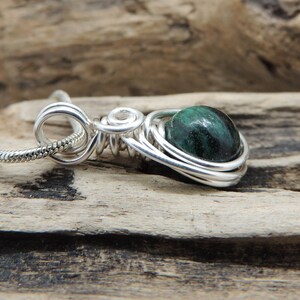Puede incluir: Un colgante de alambre de plata enrollado que presenta una piedra redonda pulida de color verde oscuro y negro. El colgante est&aacute; unido a una fina cadena de serpiente de plata, con el alambre enrollado en bucles decorativos.