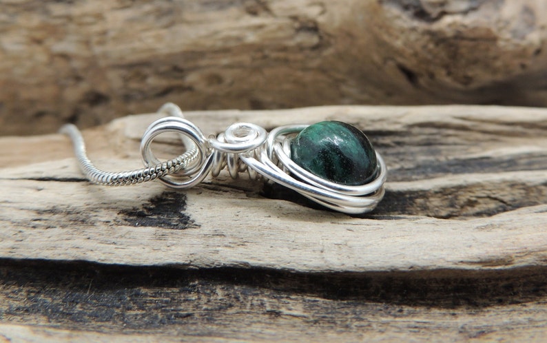 Puede incluir: Un colgante de alambre de plata enrollado que presenta una piedra redonda pulida de color verde oscuro y negro. El colgante est&aacute; unido a una fina cadena de serpiente de plata, con el alambre enrollado en bucles decorativos.