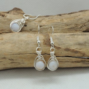 May include: A silver wire-wrapped jewellery set consisting of a pendant on a chain and a pair of matching drop earrings. Each piece features a smooth, round, pale white stone encased in intricate silver wire coils with silver ear hooks.