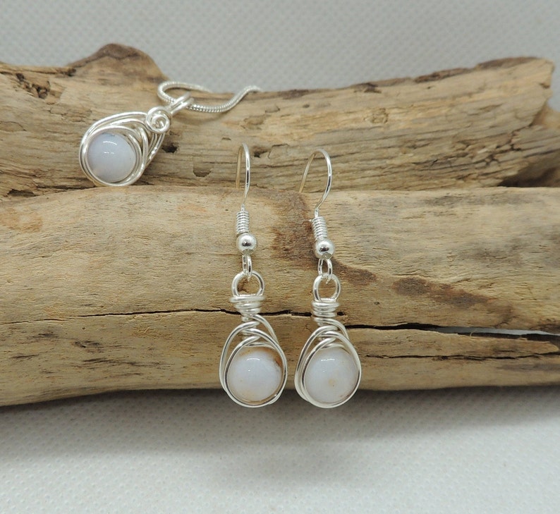 May include: A silver wire-wrapped jewellery set consisting of a pendant on a chain and a pair of matching drop earrings. Each piece features a smooth, round, pale white stone encased in intricate silver wire coils with silver ear hooks.