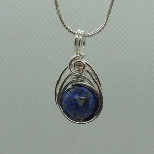 Puede incluir: Un colgante de alambre plateado que envuelve una piedra preciosa redonda de lapisl&aacute;zuli azul profundo con sutiles motas doradas. El colgante cuelga de una cadena de serpiente plateada a juego con una argolla segura en la parte superior.